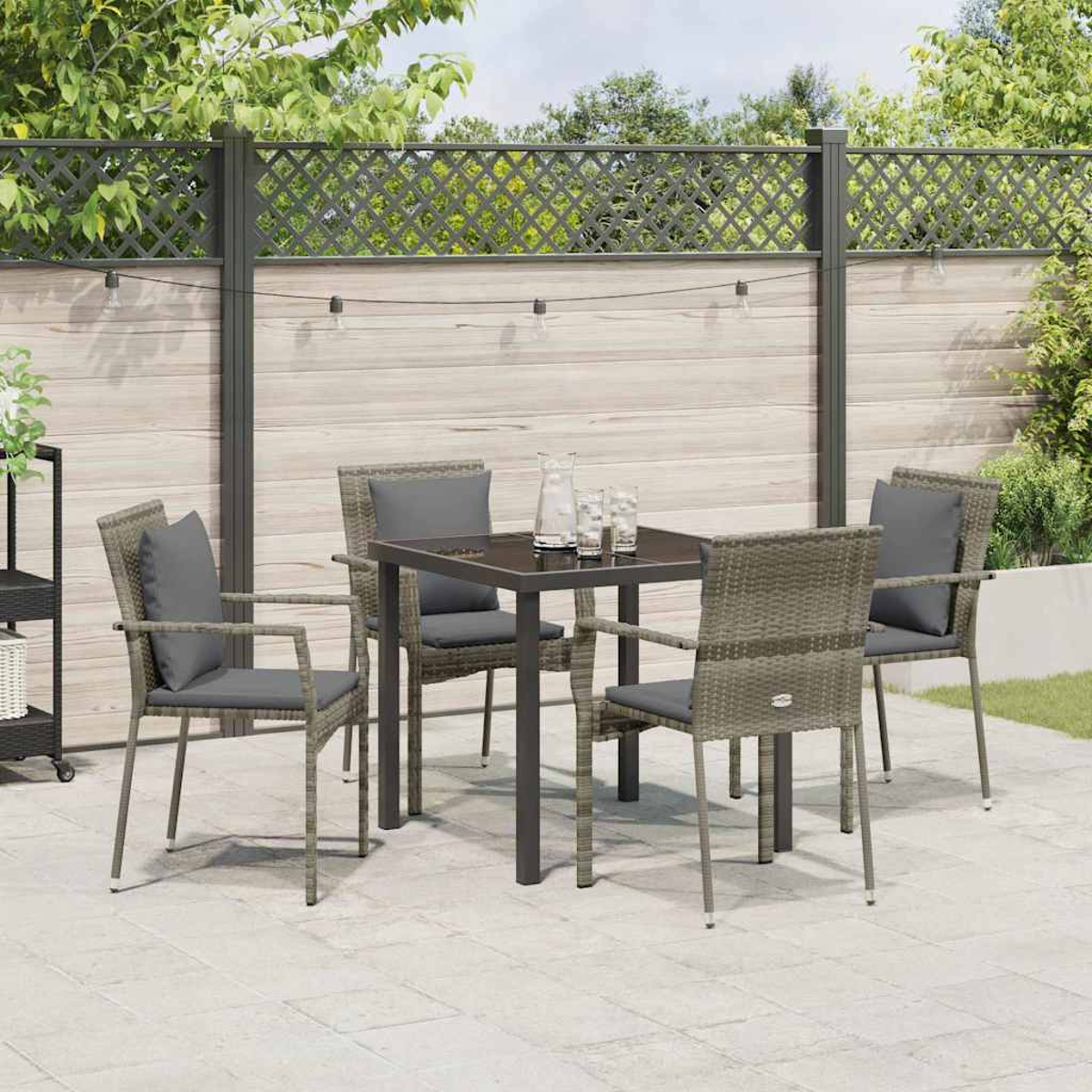 Terrassenset im Freien mit Tisch und vier Stühlen. Graue Kissen, dunkler Tisch, Holzzaun und Lichterketten. Grüne Pflanzen im Hintergrund.
