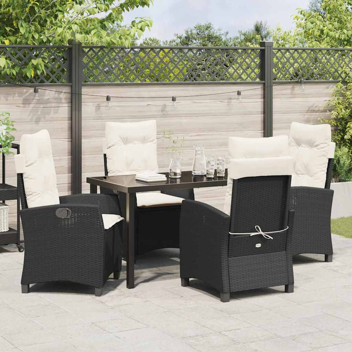 Set da patio all'aperto con sedie nere, cuscini color crema e un tavolo di vetro. Una recinzione e vegetazione sullo sfondo.