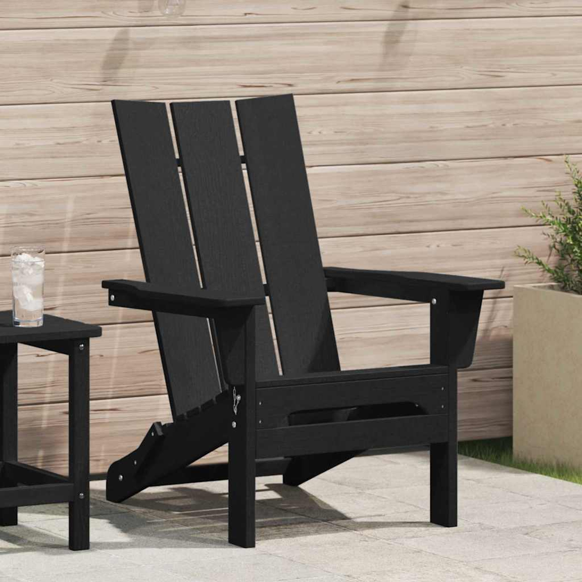 Schwarzer Adirondack-Stuhl auf der Terrasse mit Beistelltisch und einem Glas Wasser. Holzwand im Hintergrund, eine Pflanze im Topf.