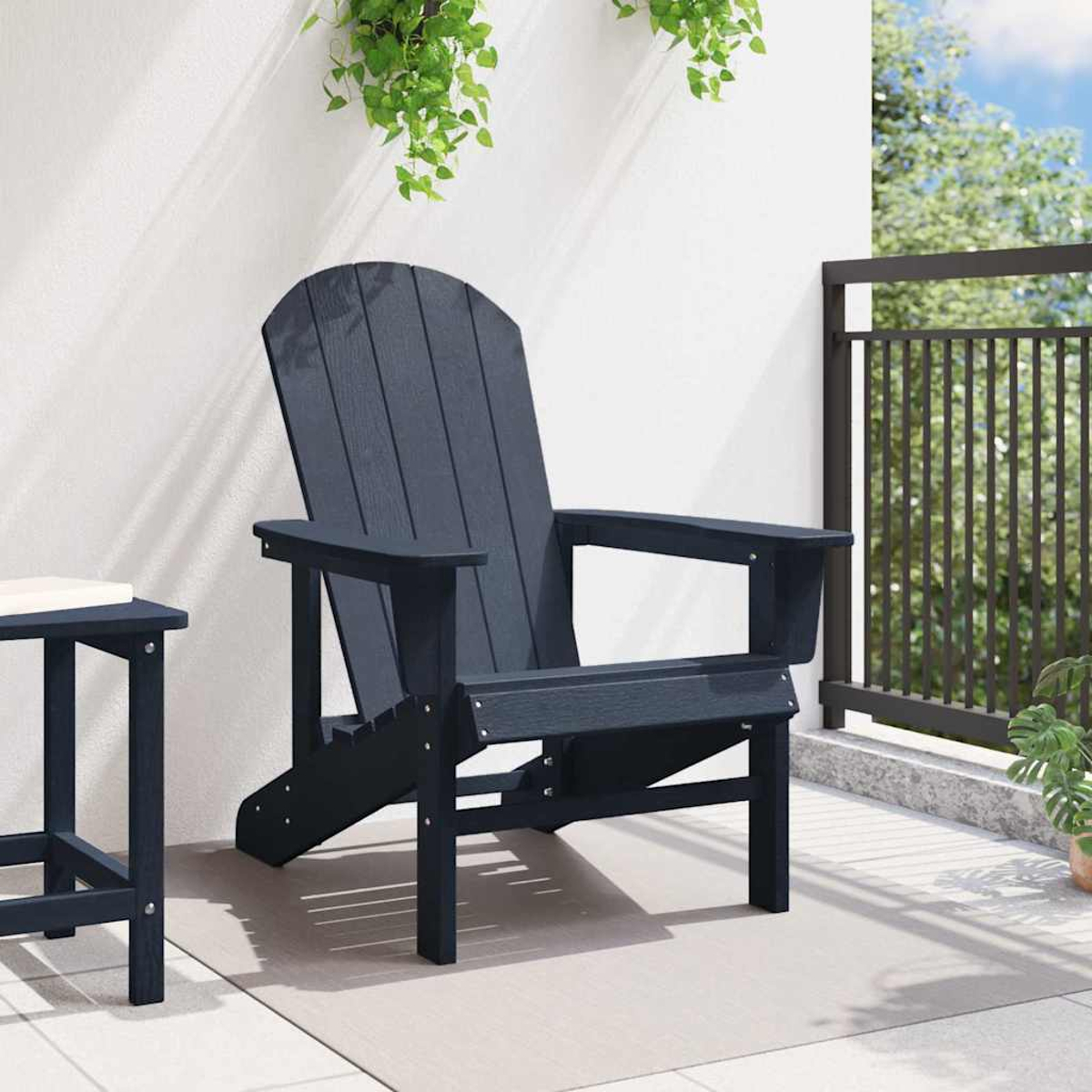 Ein dunkelblauer Adirondack-Stuhl steht auf einer Terrasse. Links steht ein kleiner Tisch. Grüne Pflanzen hängen von oben. Schwarzes Geländer im Hintergrund.