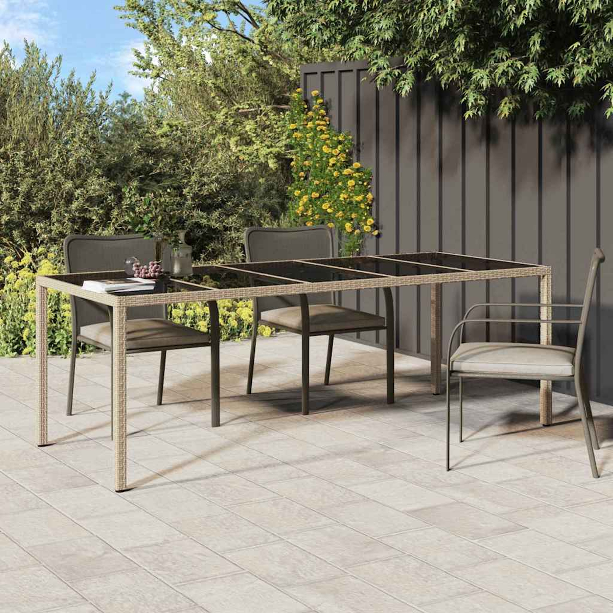 Szene auf der Terrasse mit Tisch und Stühlen. Der Tisch hat eine Glasplatte. Stühle stehen um den Tisch herum. Im Hintergrund grüne Pflanzen, Zaun und Himmel.