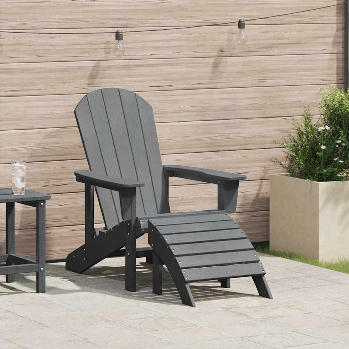 Una silla Adirondack gris con reposapiés está en un patio. Hay una mesa pequeña con un vaso de agua cerca.