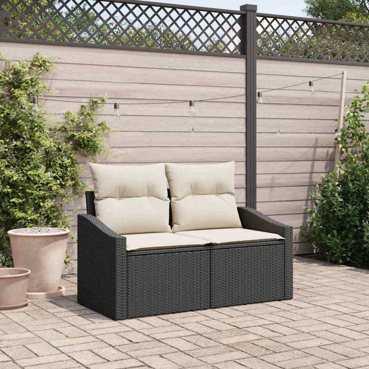 Ein dunkelgraues, gewebtes Outdoor-Sofa mit cremefarbenen Kissen steht auf einer Backstein-Terrasse. Ein Holzzaun und grüne Pflanzen sind im Hintergrund.