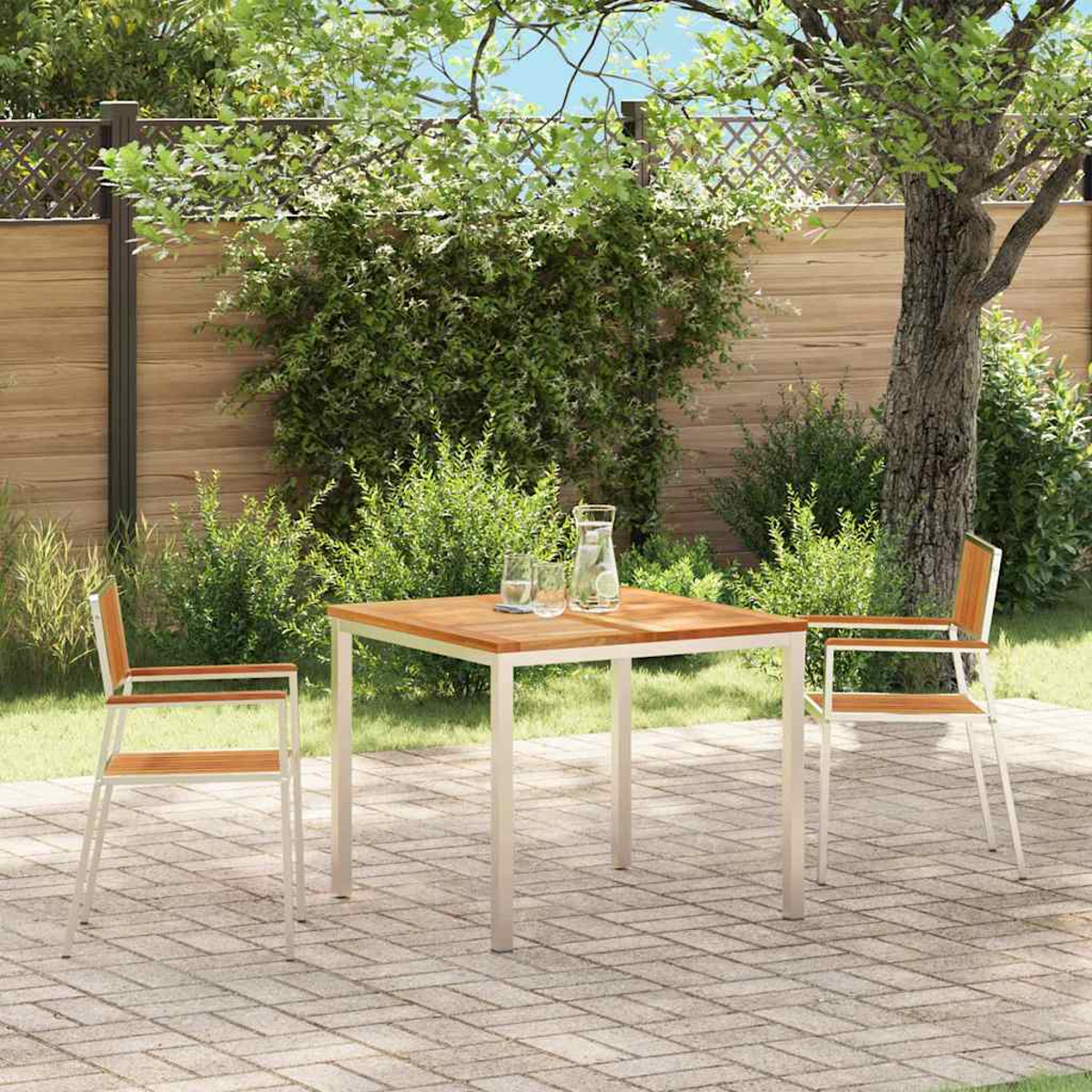Szene auf der Terrasse mit einem quadratischen Holztisch, zwei Stühlen, einer Kanne, Gläsern und üppigem Grün im Hintergrund. Holzzaun.