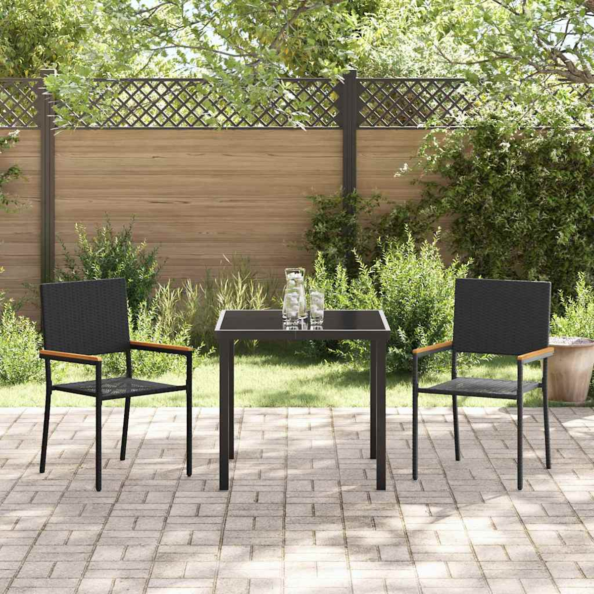 Set da patio esterno con tavolo nero e due sedie. Una recinzione di legno e vegetazione sullo sfondo. Due bicchieri e una brocca sul tavolo.