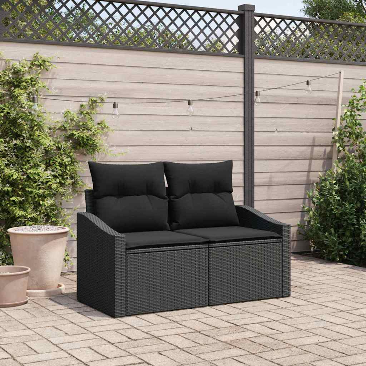 Ein schwarzes Korbmöbel mit zwei Kissen steht auf einer Ziegelterrasse, im Hintergrund ein Holzzaun und Kletterpflanzen.