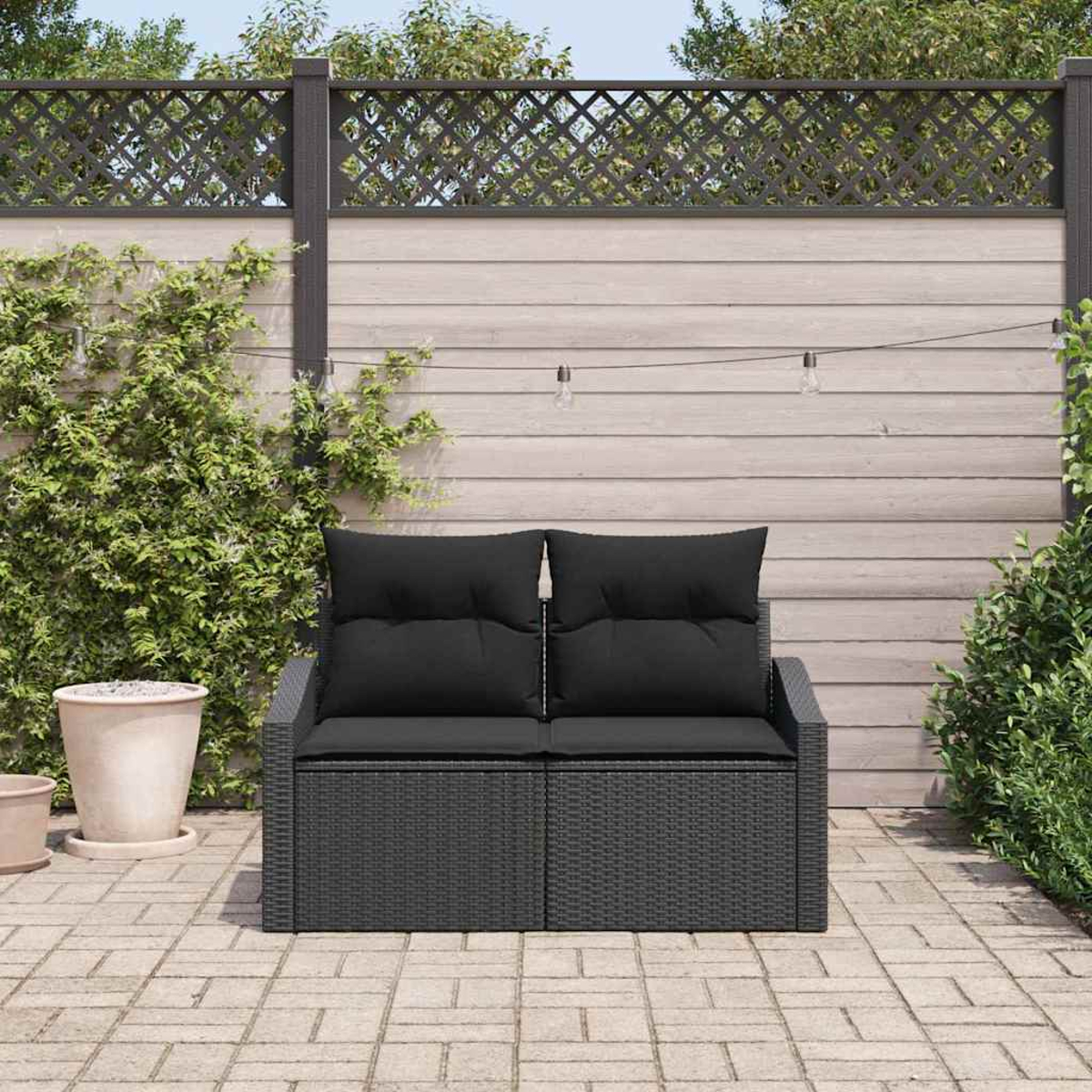 Ein schwarzes Outdoor-Sofa aus Korbgeflecht mit schwarzen Kissen steht auf einer Backstein-Terrasse. Ein Holzzaun und Grünzeug sind im Hintergrund.