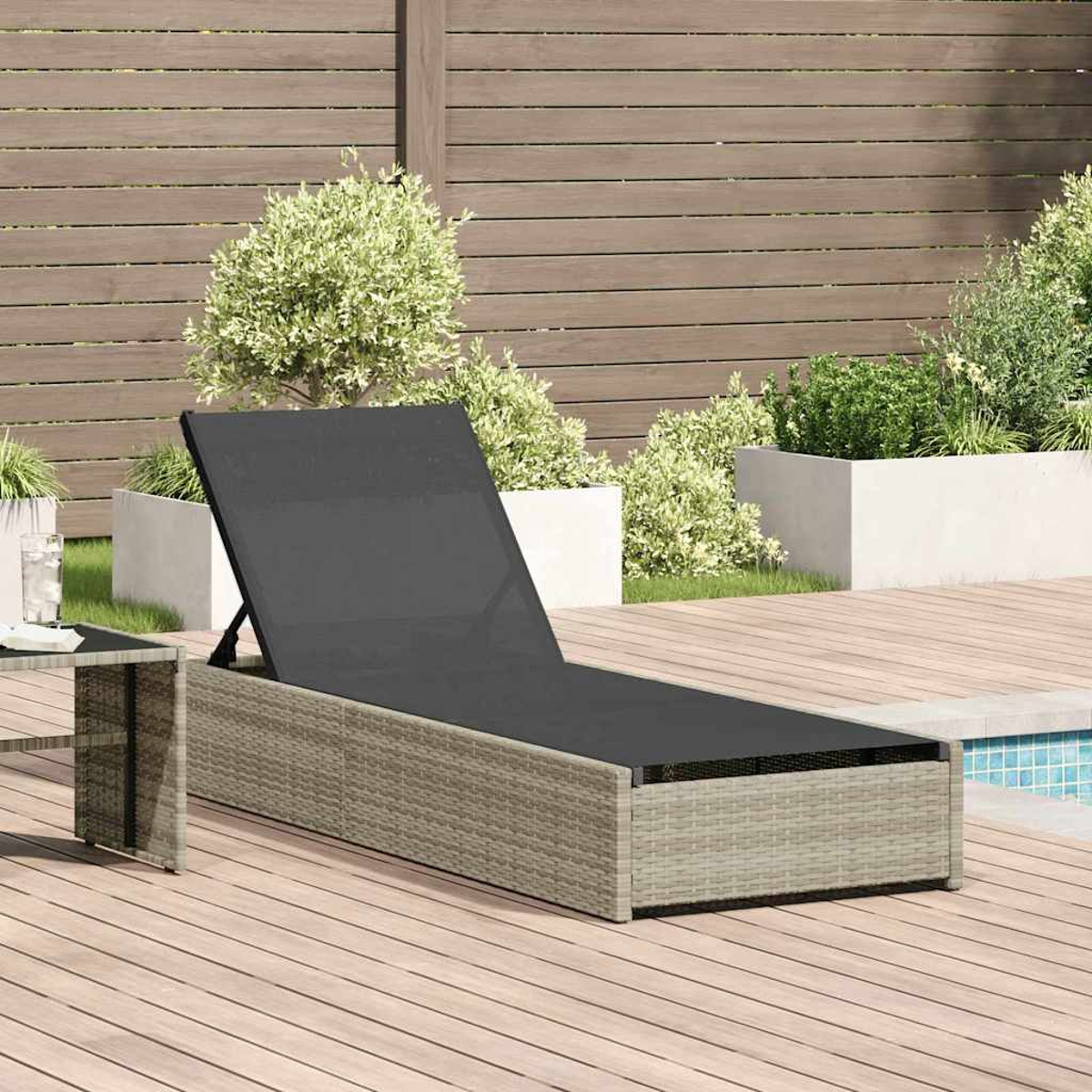 Eine graue Rattan-Liege steht auf einem Holzdeck. Dahinter befinden sich Pflanzen, ein Pool und ein Holzzaun.
