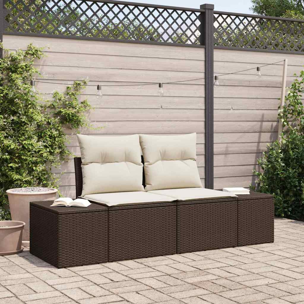 Ein braunes Korbmöbel mit beigen Kissen steht auf einer Terrasse. Ein Zaun mit Lichtern hängt darüber. Pflanzen und Töpfe sind herum.