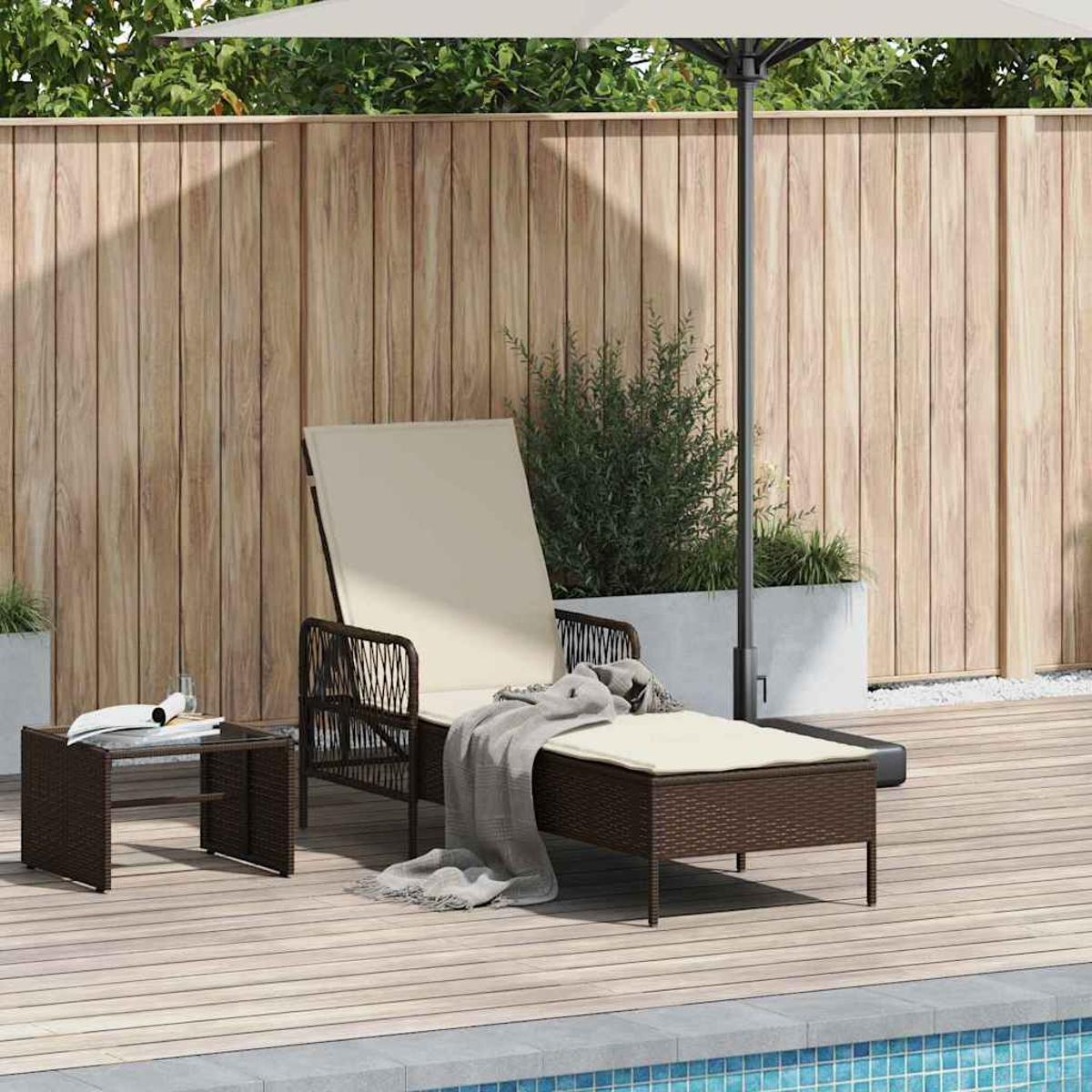 Eine Terrasse mit einer braunen Korbliege mit cremefarbenem Kissen, einem kleinen Tisch und einem Sonnenschirm. Im Hintergrund ist ein Holzzaun zu sehen.