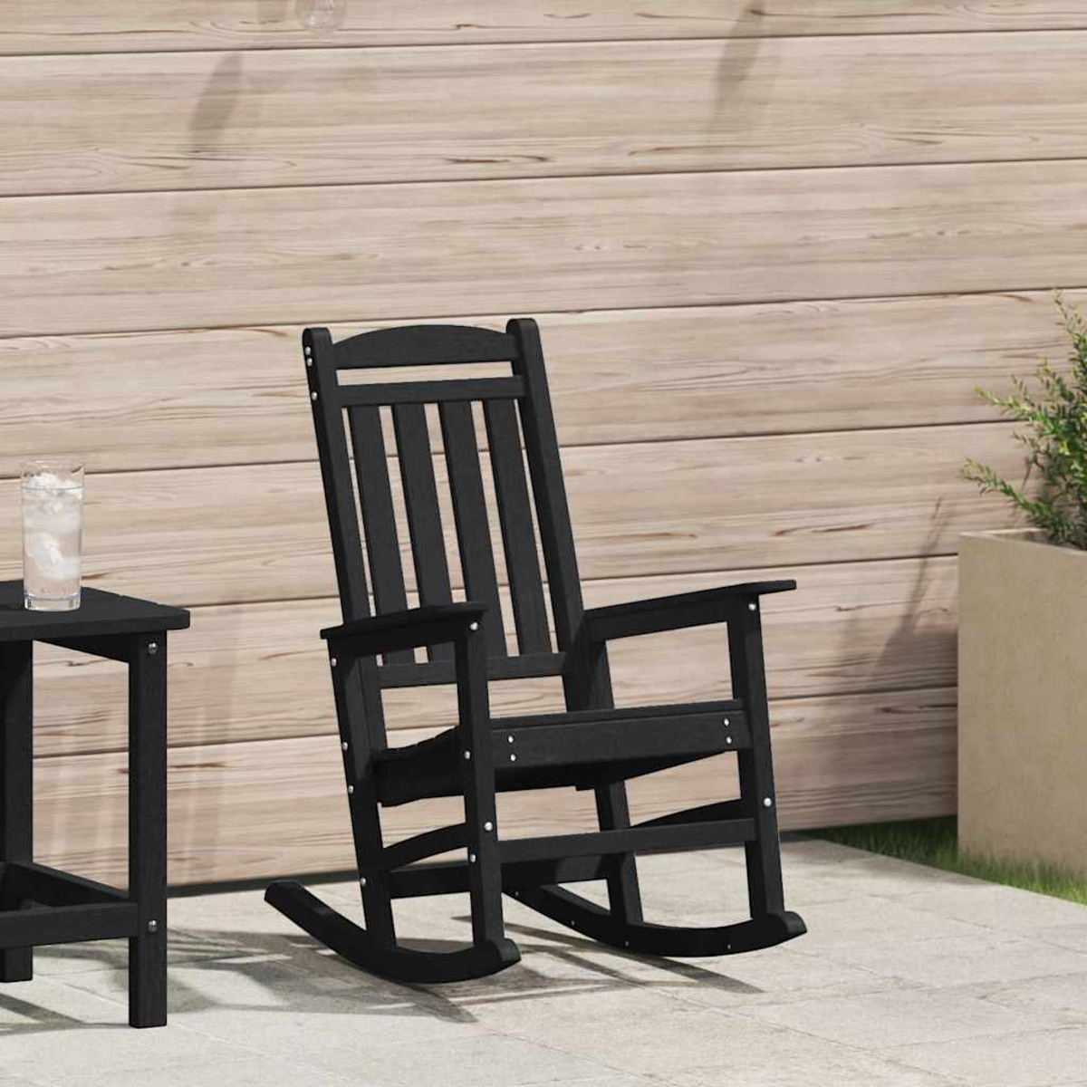 Sillón mecedor negro en un patio. Una pequeña mesa con un vaso de agua está a la izquierda. Fondo de paneles de madera.