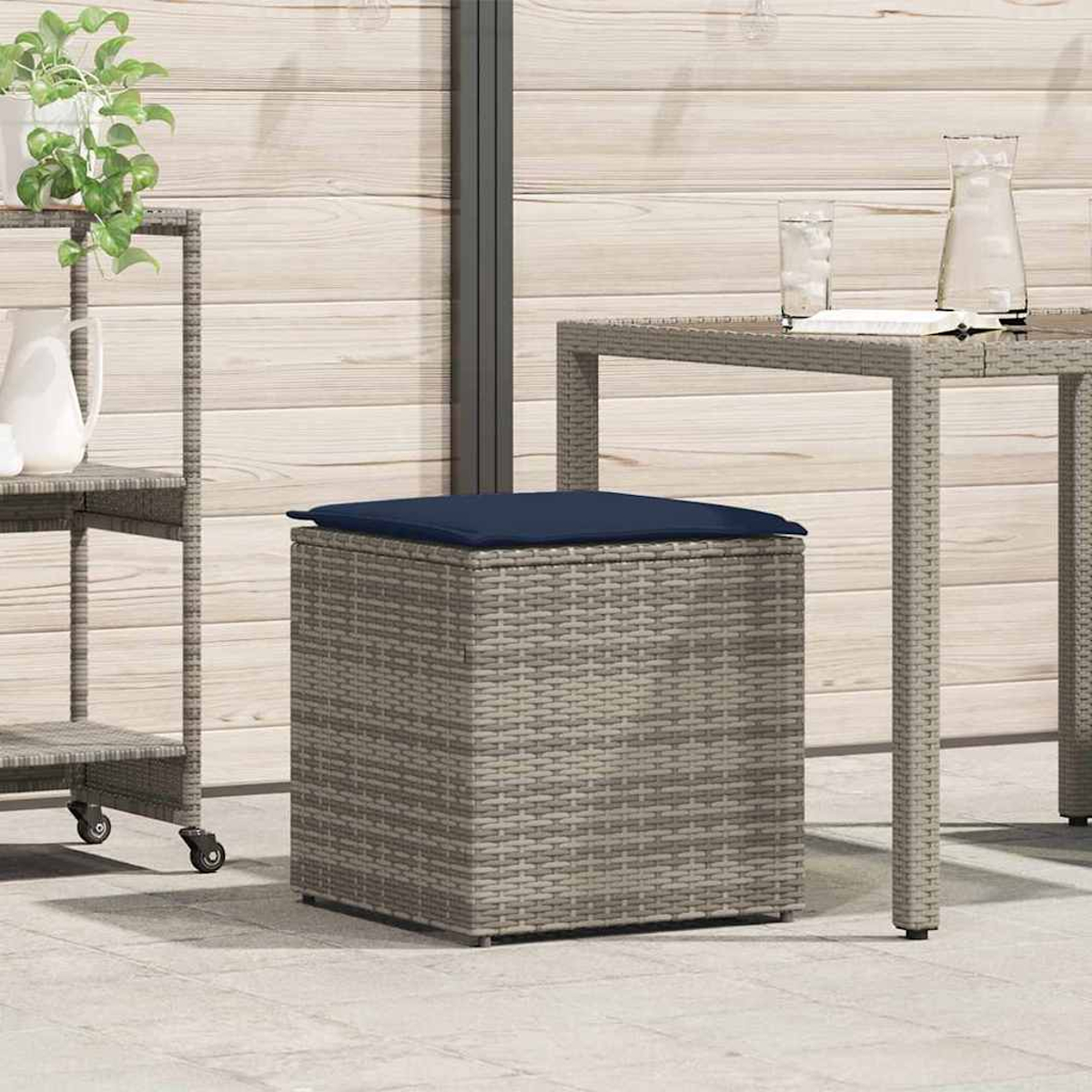 Ein Korbflechterwürfel mit blauem Kissen steht auf einer Terrasse. Ein Tisch mit Gläsern und einem Krug ist in der Nähe. Links ist ein Rollwagen.