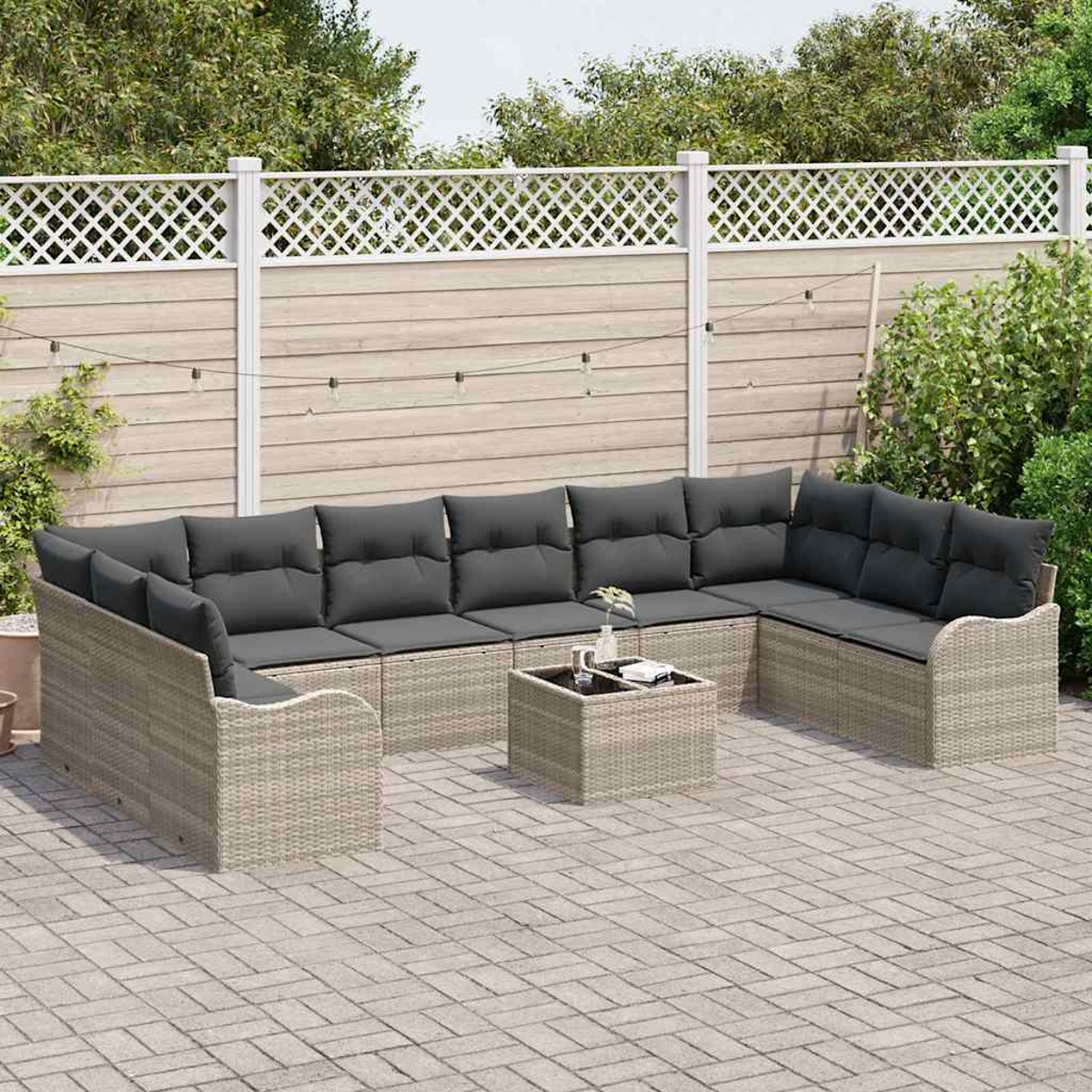 Gartenmöbel-Set mit grauen Kissen. Korbsessel und Couchtisch auf einer Stein-Terrasse. Weißer Zaun und Grün im Hintergrund.