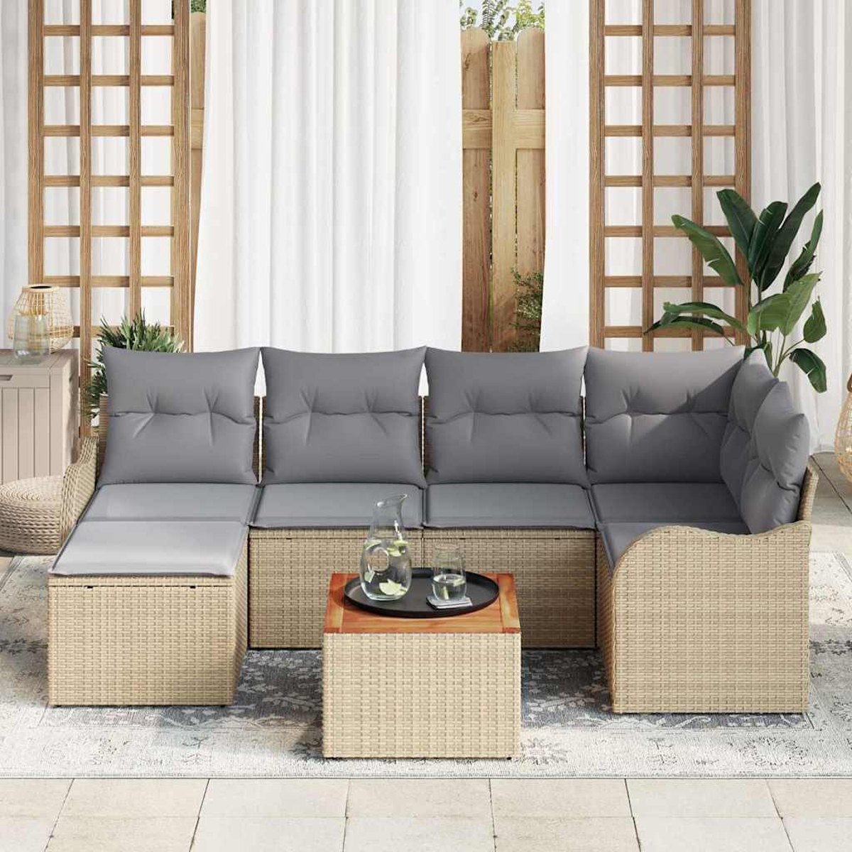 Ein beiges Korbsessel-Ecksofa mit grauen Kissen. Ein kleiner Holztisch trägt ein Tablett mit Krug und Gläsern. Weiße Vorhänge im Hintergrund.