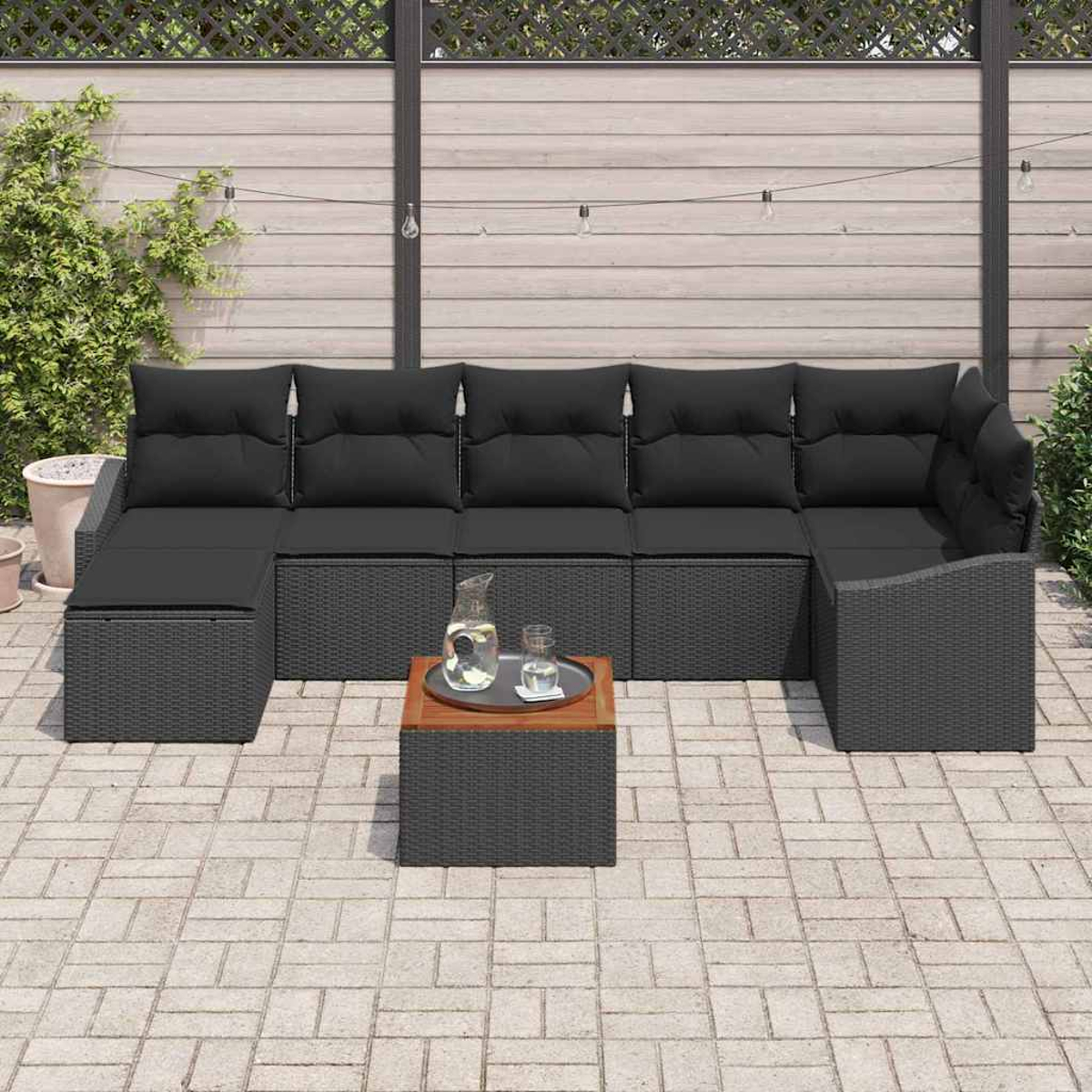 Outdoor-Sitzgruppe mit schwarzen Kissen und Korbgeweben. Inklusive kleinem Tisch mit Krug und Gläsern. Auf Ziegelpflaster.