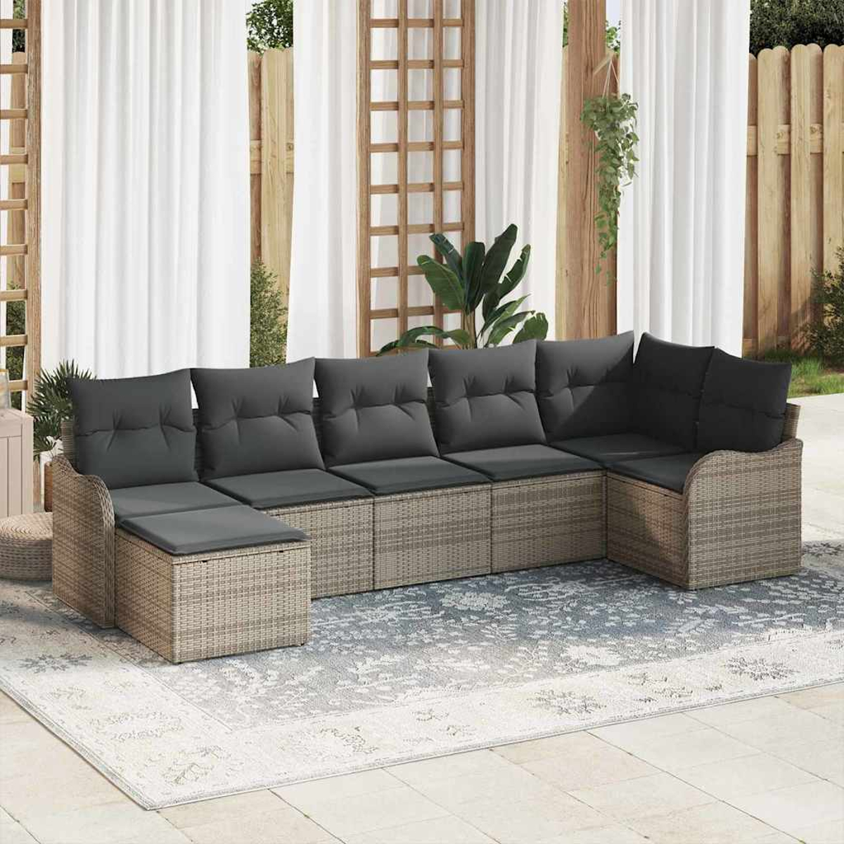 Ein Ecksofa mit grauen Kissen steht auf einem gemusterten Teppich. Dahinter befinden sich weiße Vorhänge und ein Holzzaun.