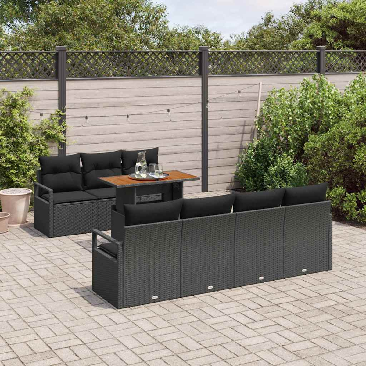 Terrassengarnitur mit grauen Korbmöbeln, schwarzen Kissen und einem Holztisch. Auf einer Backsteinterrasse mit Zaun und Grün.