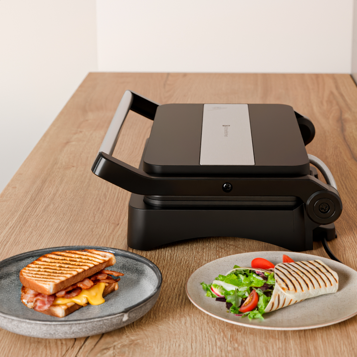 A sandwich grill with two grilled sandwiches and a wrap with salad and tomatoes on a wooden table.