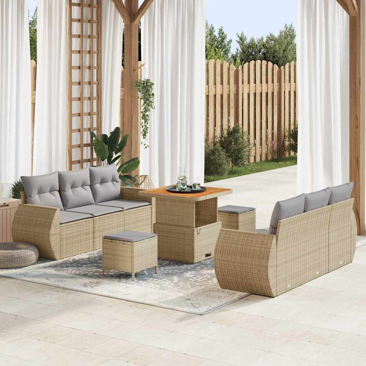 Juego de patio con sofá, mesa y otomanas de mimbre. Cortinas blancas y valla de madera al fondo.