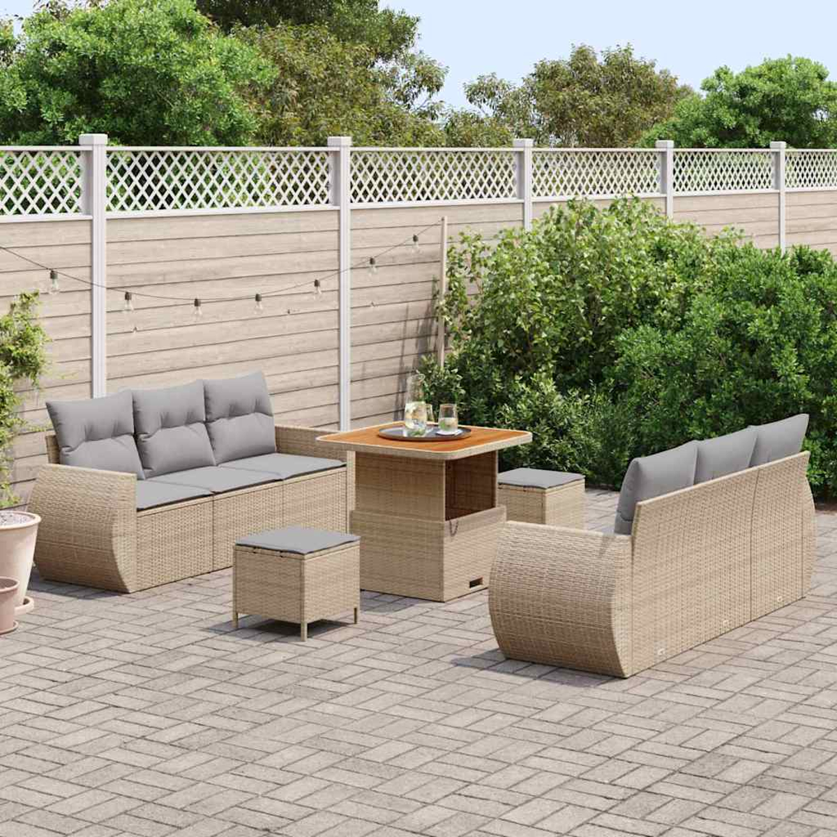 Juego de muebles de mimbre para exteriores en un patio pavimentado. Incluye dos sofás, dos otomanas y una mesa, con una valla y vegetación de fondo.