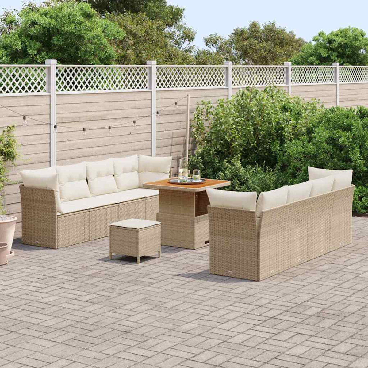 Garten-Sitzgruppe. Beige Korbsessel, Tisch und Hocker auf einer Backstein-Terrasse. Weißer Gitterzaun und grünes Laub im Hintergrund.