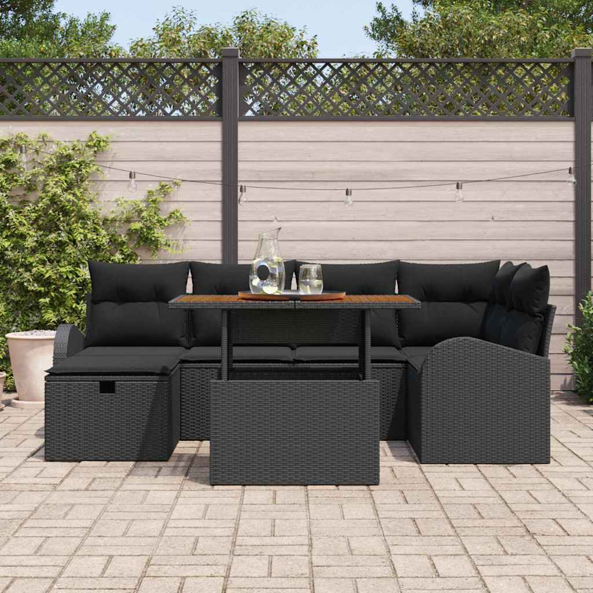 Outdoor-Sofa-Set aus schwarzem Korbgeflecht mit Kissen, Couchtisch und Wasserkaraffe auf einer Terrasse. Zaun und Grün im Hintergrund.