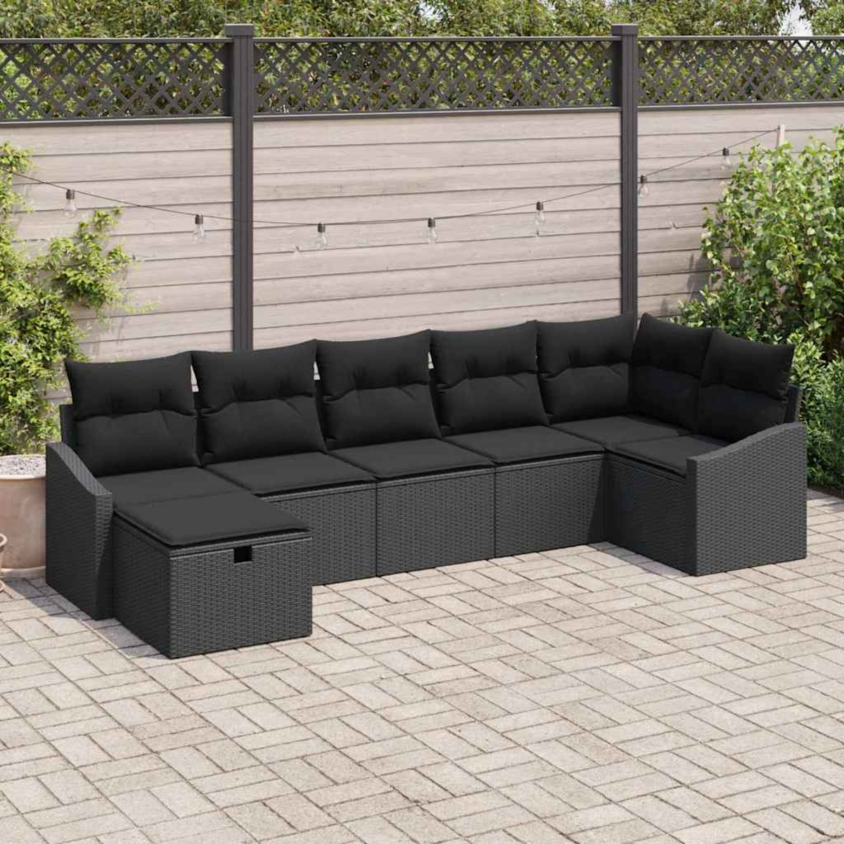 Ein graues Outdoor-Ecksofa mit schwarzen Kissen steht auf einer Backstein-Terrasse. Im Hintergrund befinden sich ein Holzzaun und Grün.