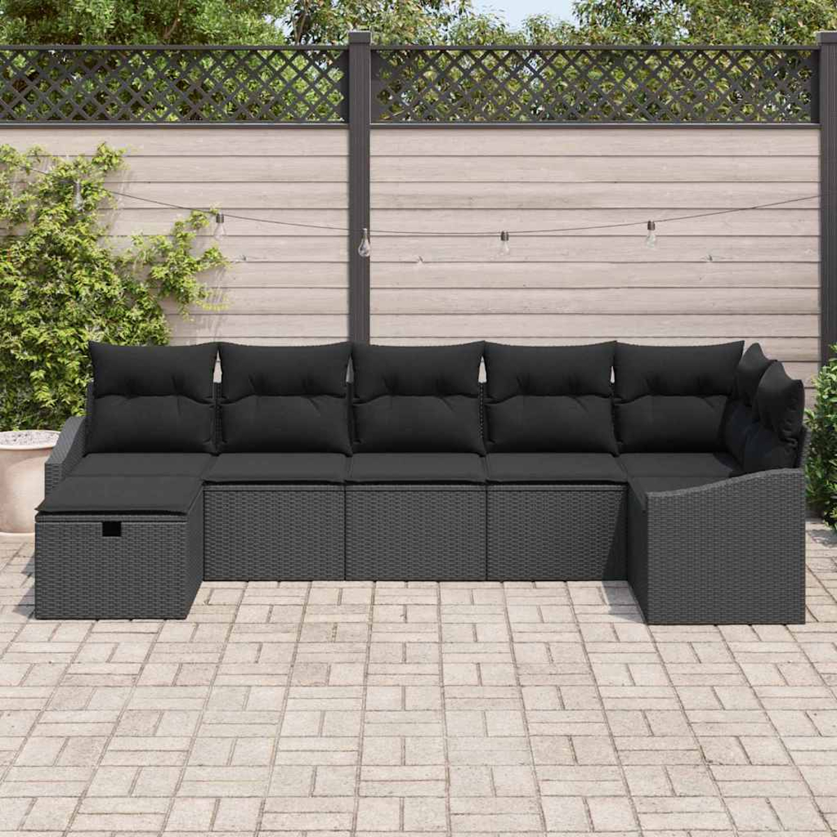 Graues Rattan-Ecksofa mit schwarzen Kissen auf einer Steinterrasse. Holzzaun und Grün bilden den Hintergrund.