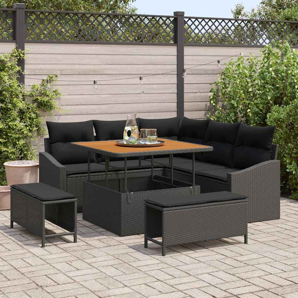 Gartenmöbel-Set mit schwarzen Kissen, Holztisch und zwei Bänken. Im Hintergrund ein Zaun und Grün.