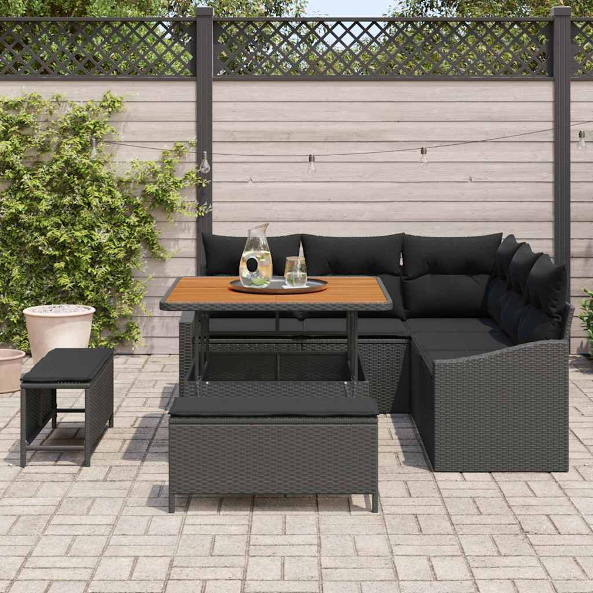 Terrassenset mit schwarzen Kissen, Holztisch und grauen Korbmöbeln. Eine Kanne und Gläser stehen auf dem Tisch.