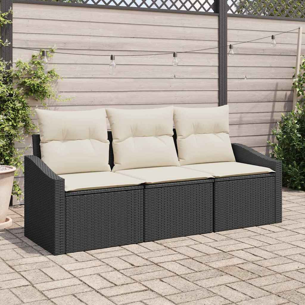 Ein dunkelgraues Korbmöbelsofa mit cremefarbenen Kissen steht auf einer Backstein-Terrasse. Im Hintergrund befinden sich ein Holzzaun und Hängelampen.