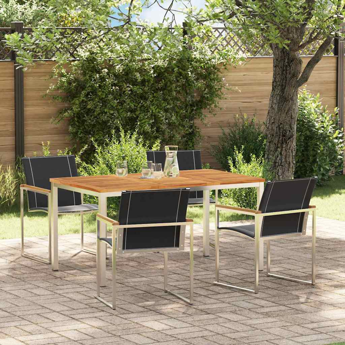 Juego de comedor al aire libre con mesa de madera, sillas negras y vasos en un patio de ladrillos. Follaje verde y una valla de madera al fondo.