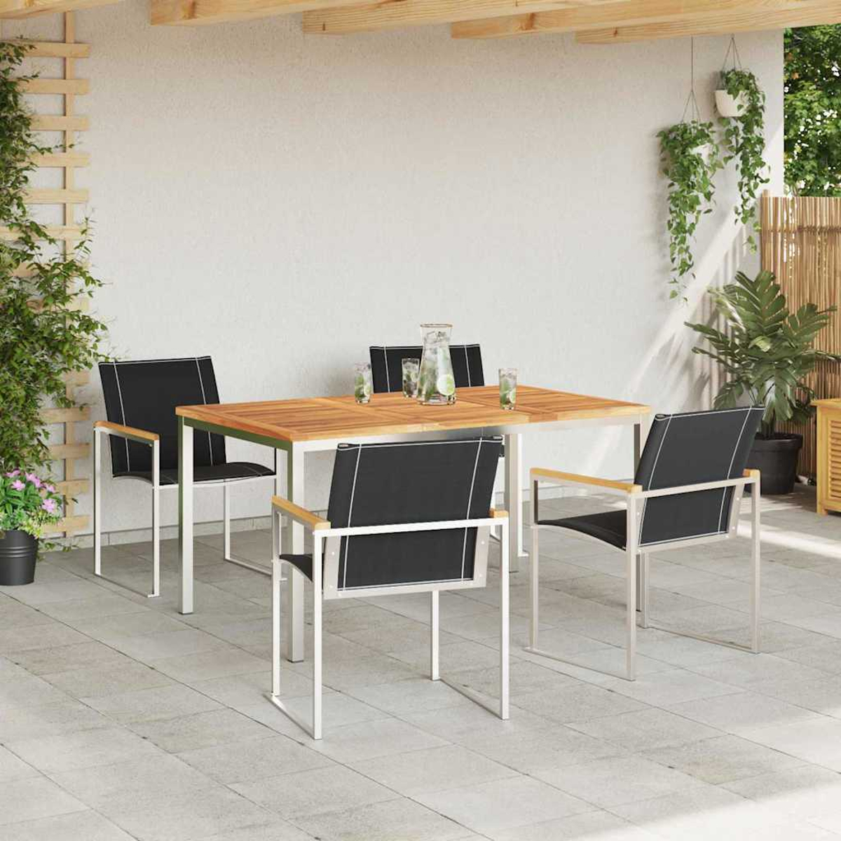 Juego de comedor al aire libre con mesa de madera, cuatro sillas y vasos de agua. Plantas y pared blanca al fondo.
