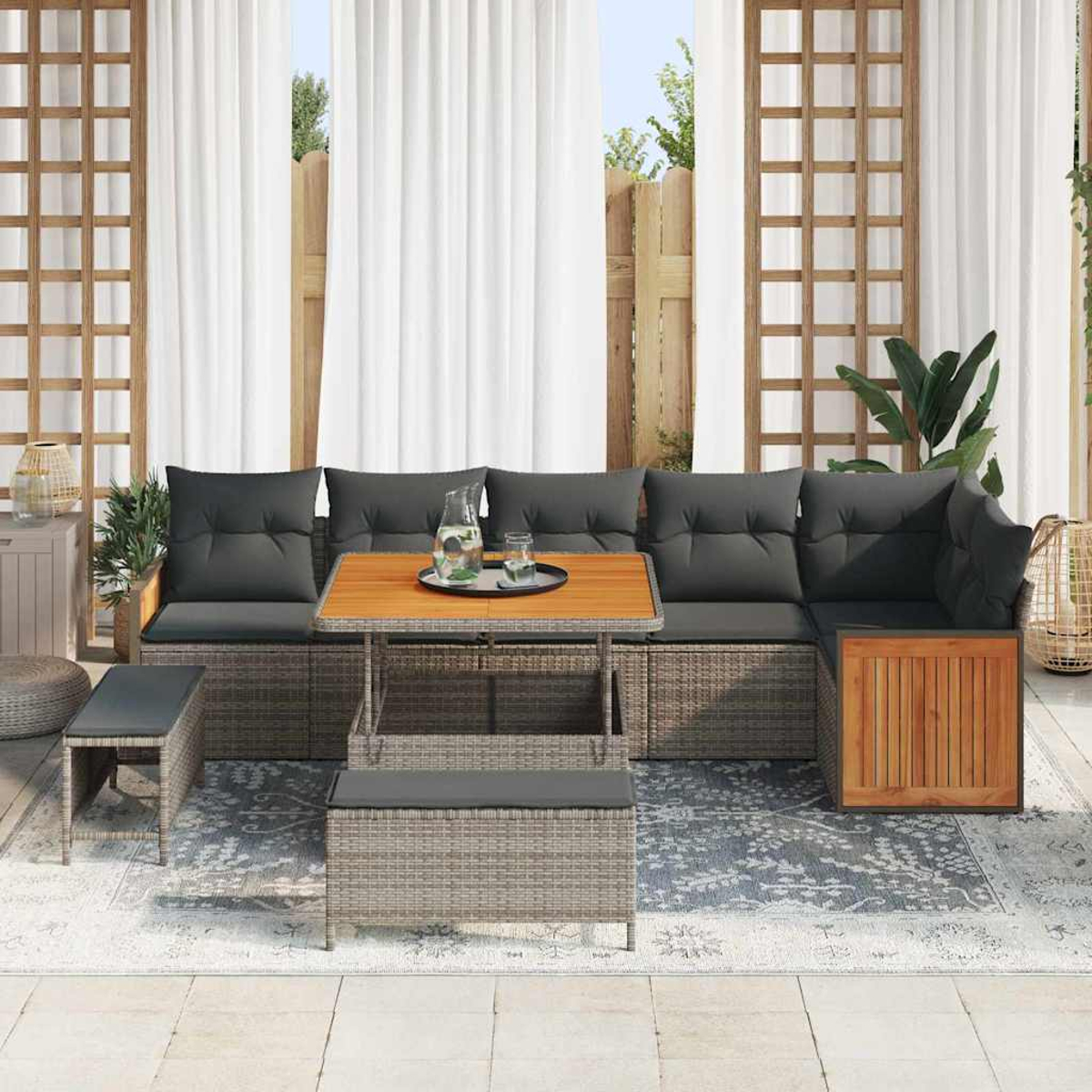 Gartensofa mit grauen Kissen, Holztisch und Sitzbank auf gemustertem Teppich. Weiße Vorhänge und Holzzaun im Hintergrund.