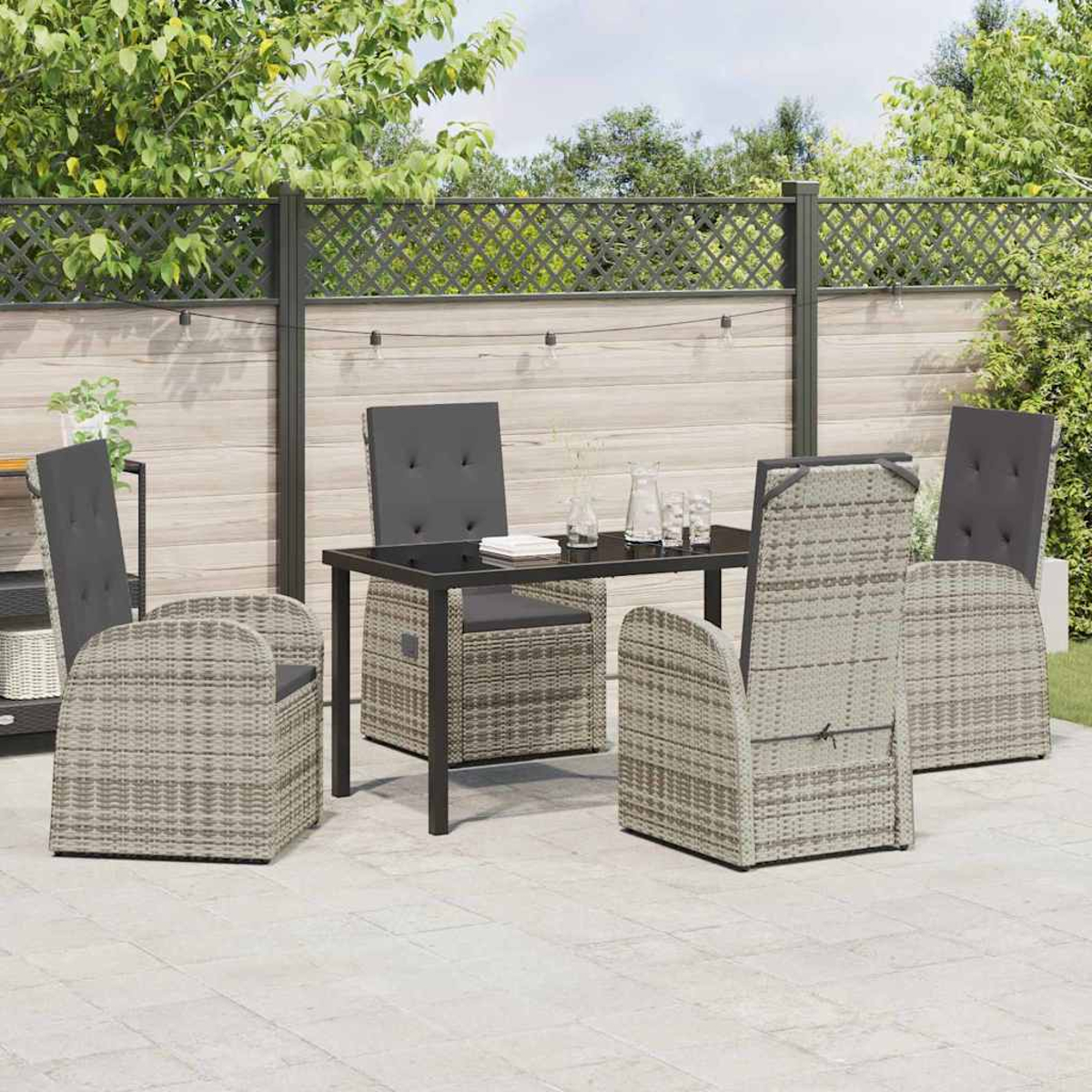 Set da pranzo all'aperto: tavolo, quattro sedie in vimini con cuscini grigi. Su un patio in pietra, con recinzione in legno e verde.