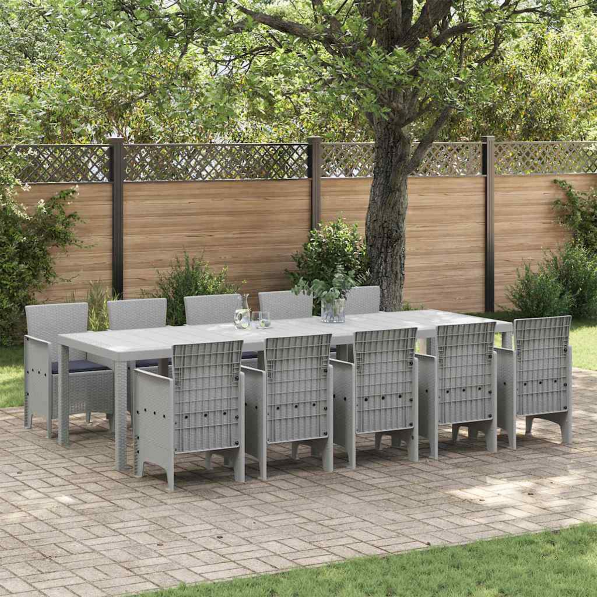 Ein graues Ess-Set für den Außenbereich mit einem langen Tisch und acht Stühlen steht auf einer Ziegelterrasse. Im Hintergrund ein Holzzaun und ein Baum.