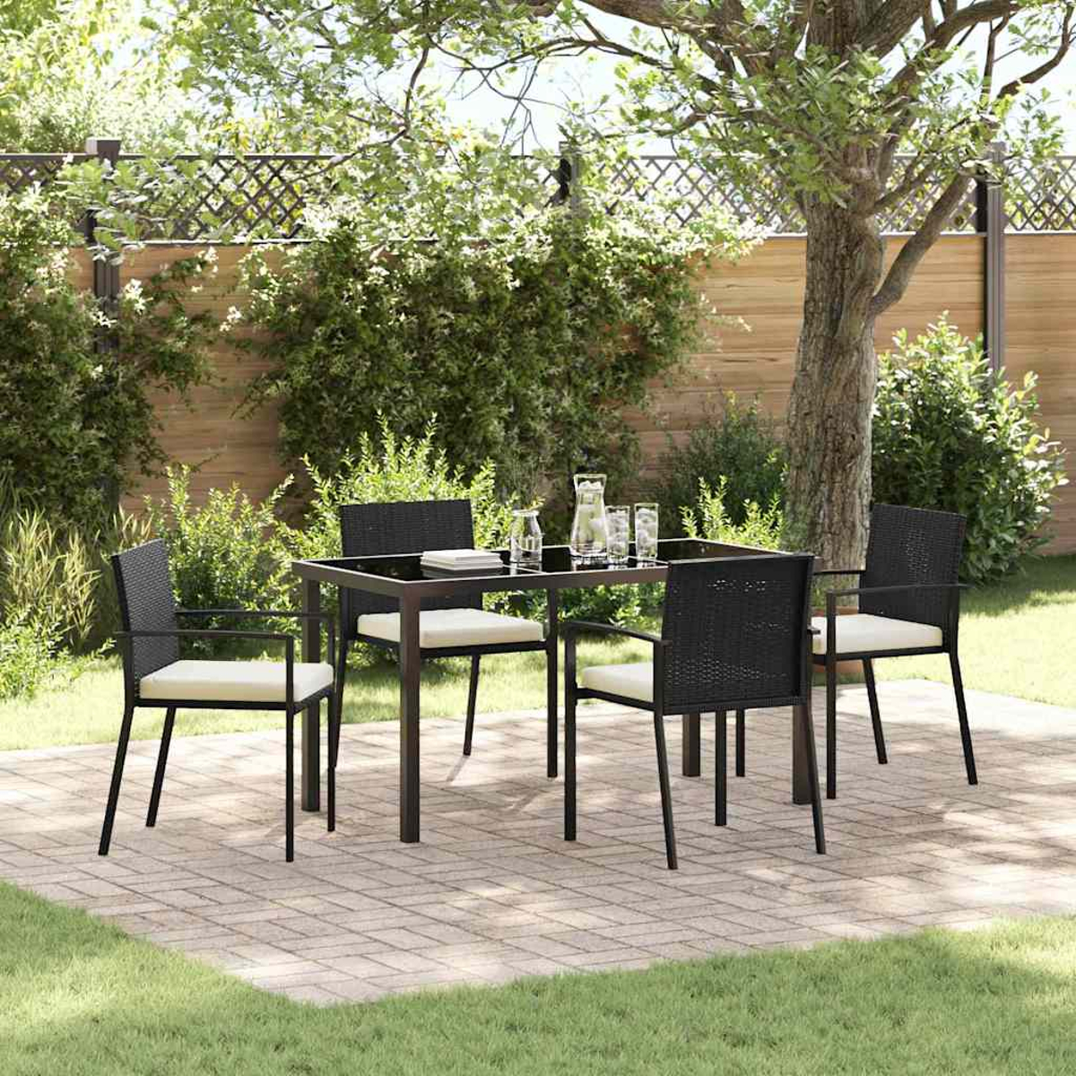 Essgruppe im Freien mit schwarzem Tisch, vier Stühlen und Krug. Auf einer Ziegelterrasse mit Baum- und Zaunhintergrund.