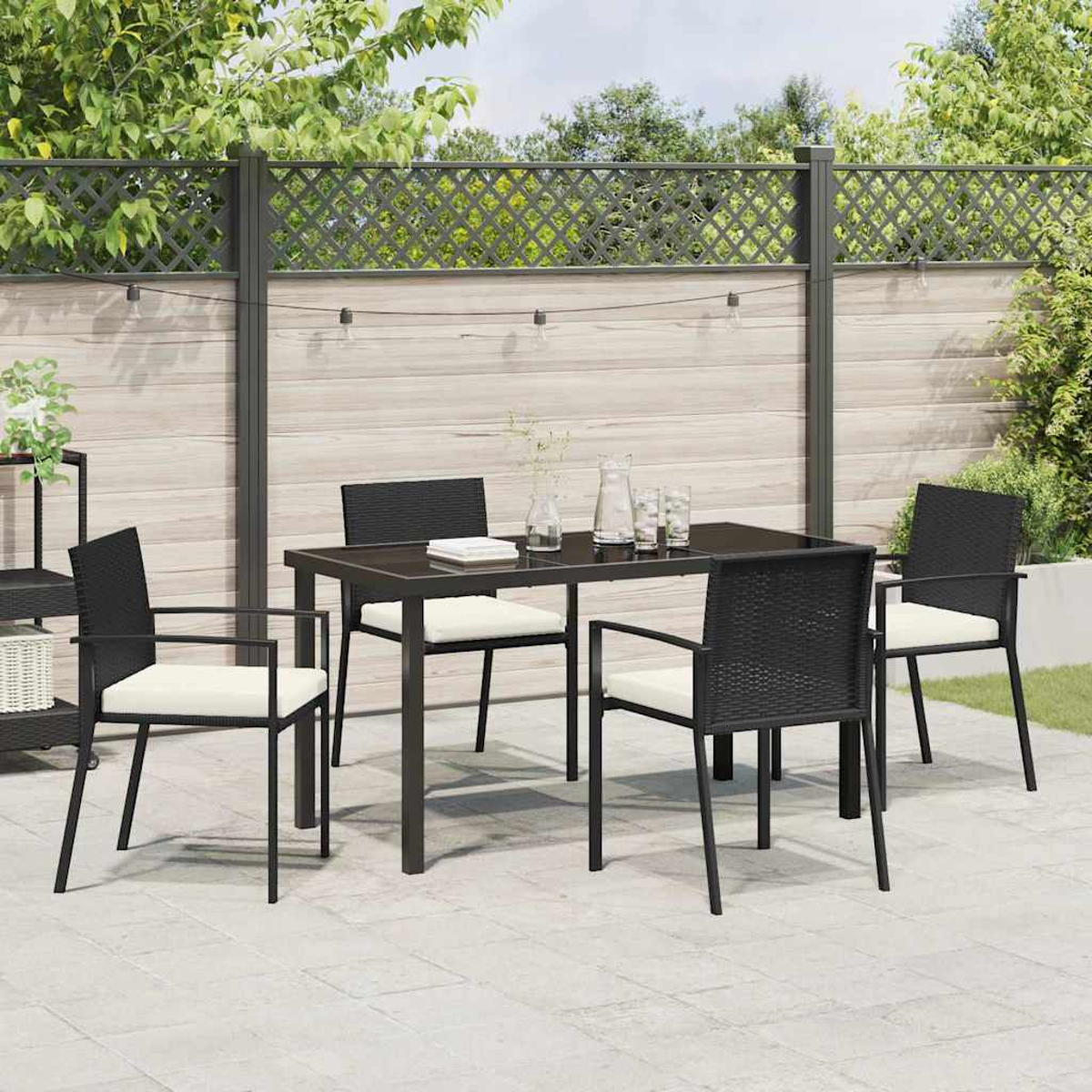 Essgruppe im Freien: schwarzer Tisch und Stühle auf der Terrasse. Hintergrund: Zaun, Grün und Lichterkette.