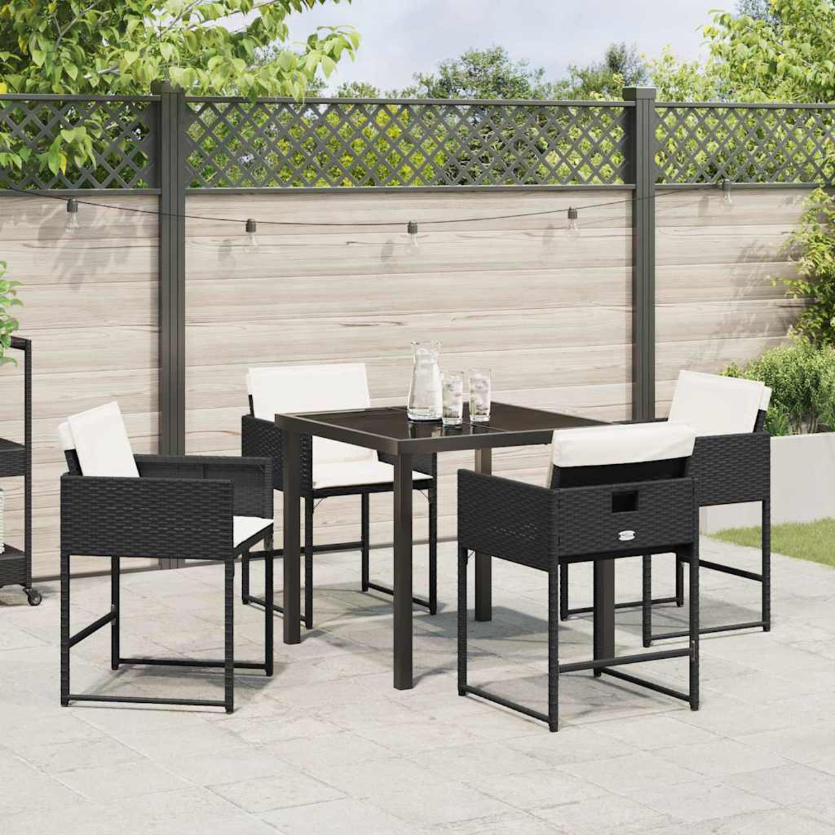 Ein schwarzes Ess-Set für den Außenbereich mit einem quadratischen Tisch und vier Stühlen mit weißen Kissen. Im Hintergrund ein Holzzaun und Grün.