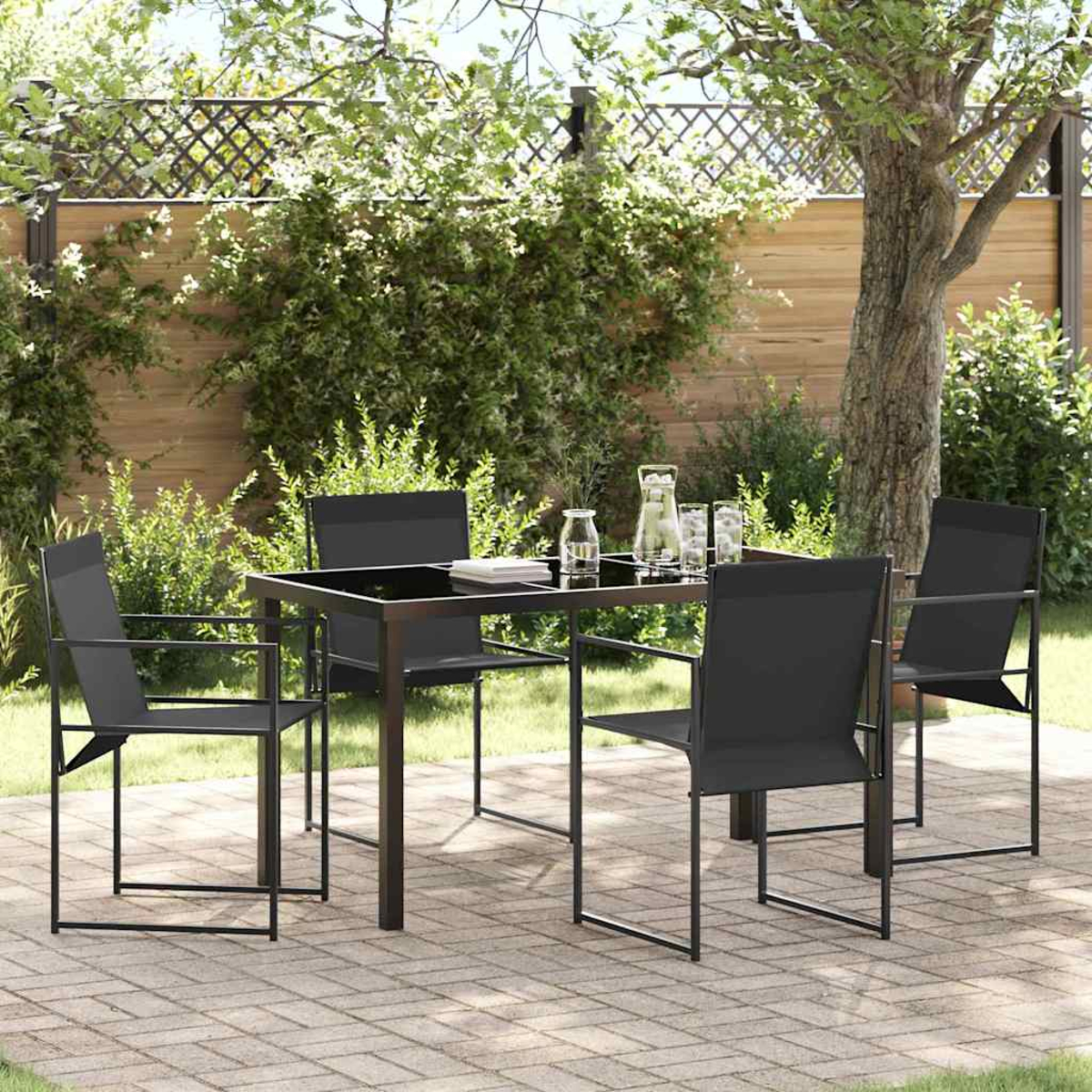 Essgarnitur im Freien mit schwarzem Tisch und Stühlen. Ein Baum und Grün bilden den Hintergrund, auf einer Terrasse aus Ziegeln.