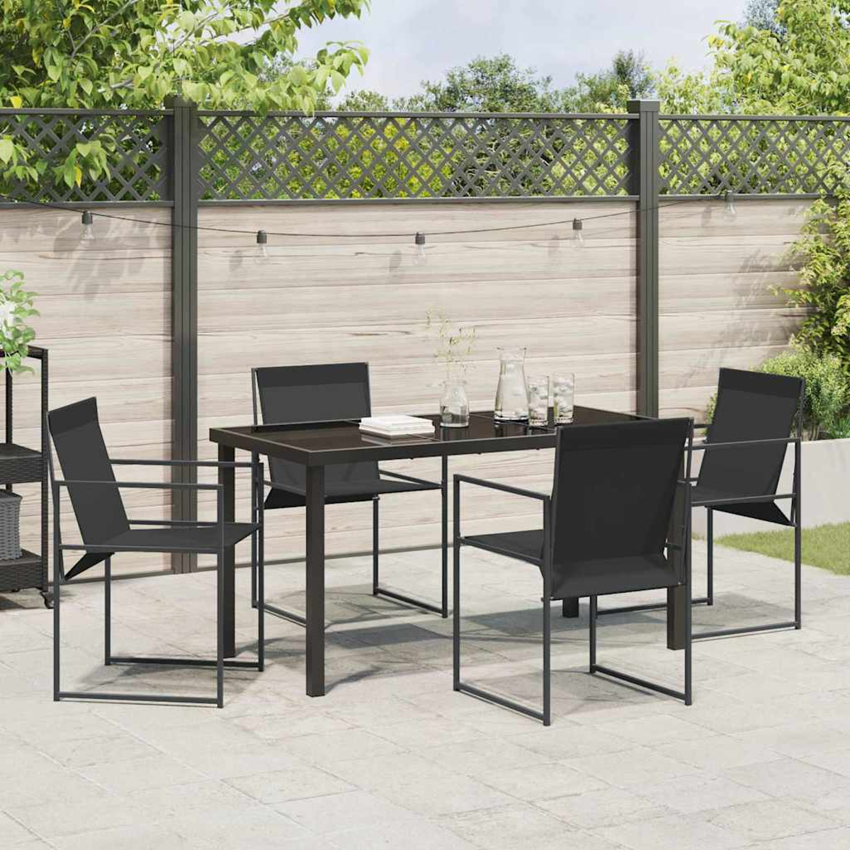 Essgruppe im Freien: schwarzer Tisch und Stühle auf der Terrasse. Hintergrund: Zaun, Pflanzen und Lichterkette.
