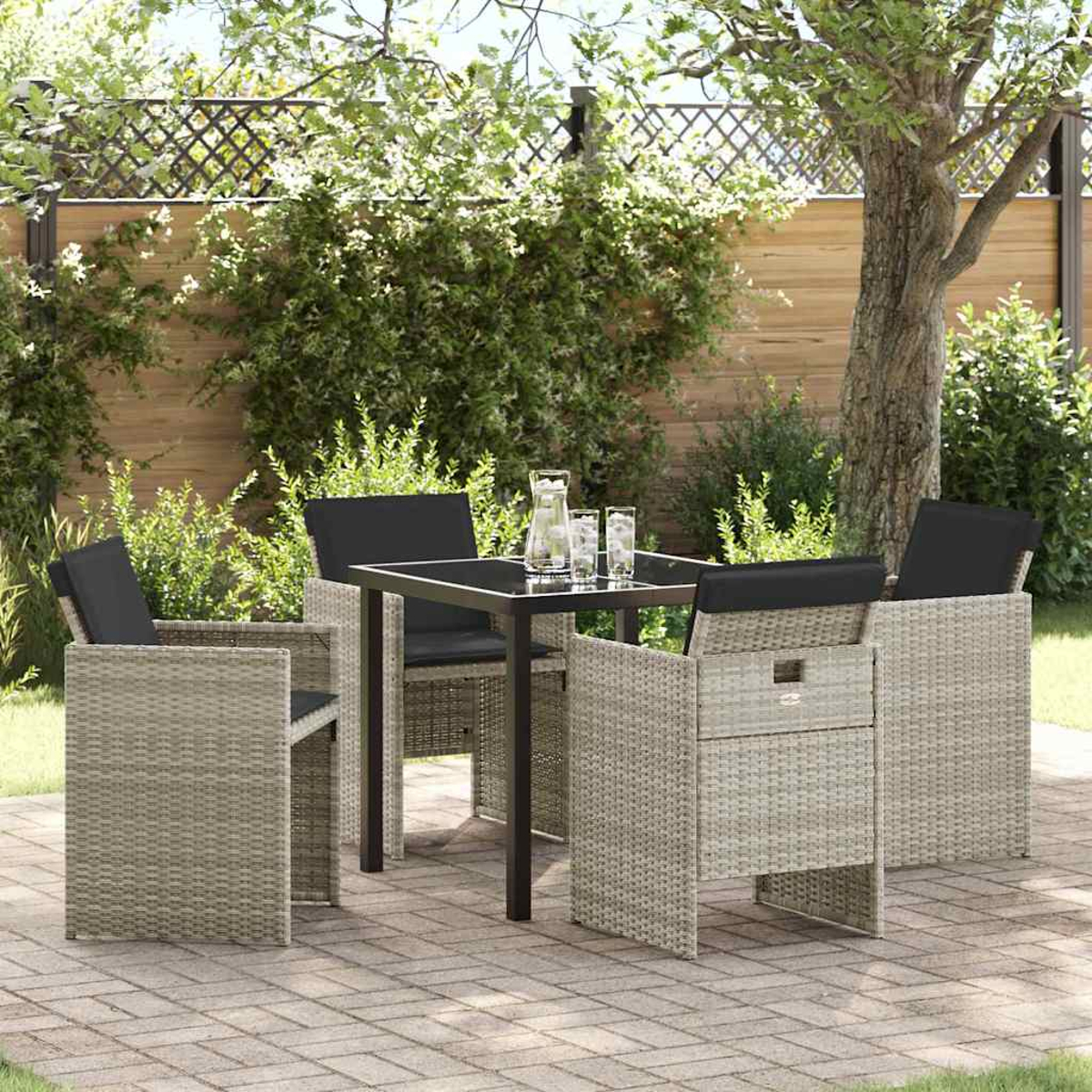 Set da patio esterno con sedie in vimini grigie, cuscini neri e un tavolo quadrato. Una brocca e bicchieri sul tavolo. Sfondo con vegetazione.