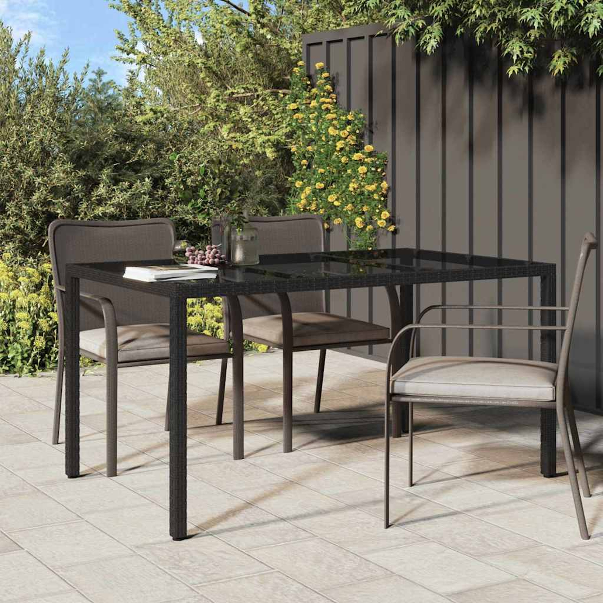 Essgruppe im Freien: schwarzer Tisch, vier Stühle, Trauben und eine Vase auf einer gefliesten Terrasse. Grüne Pflanzen und Holzzaun im Hintergrund.