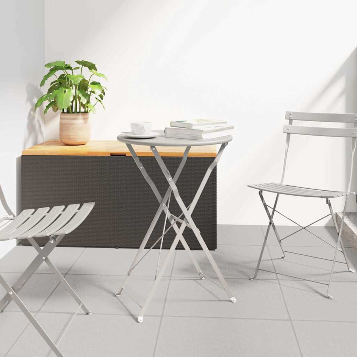 Ein grauer Klapptisch und Stühle auf einer Terrasse. Auf dem Tisch stehen eine Tasse und Bücher. Im Hintergrund stehen eine Pflanze im Topf und eine Aufbewahrungsbox.