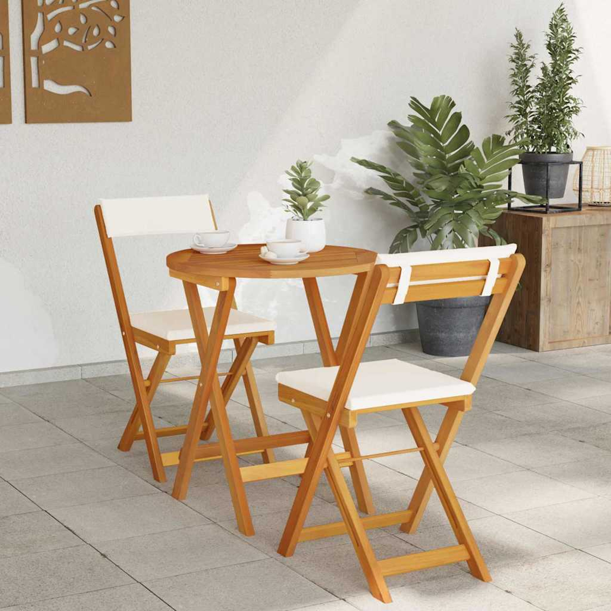 Una mesa de madera y dos sillas plegables con cojines blancos están en un patio. Plantas y objetos decorativos al fondo.
