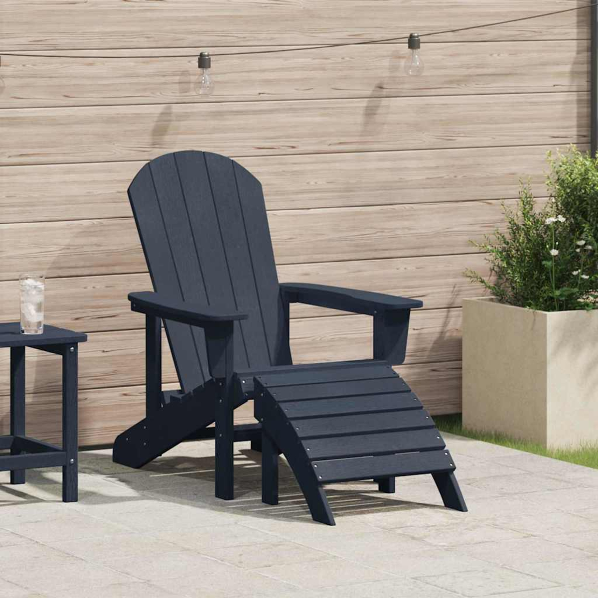 Ein marineblauer Adirondack-Stuhl mit Fußstütze steht auf einer Terrasse. Ein kleiner Tisch und eine Pflanze sind ebenfalls vorhanden. Holzwand im Hintergrund.