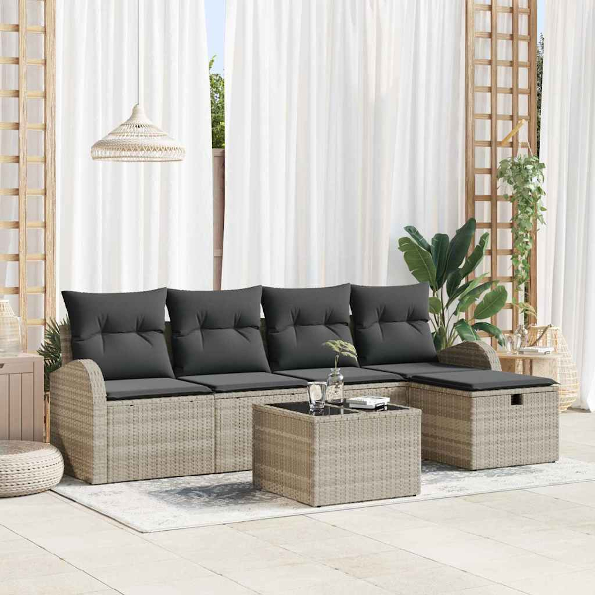Outdoor-Rattan-Sofagarnitur mit grauen Kissen, einem Couchtisch und einem Hocker. Weiße Vorhänge und Pflanzen im Hintergrund.