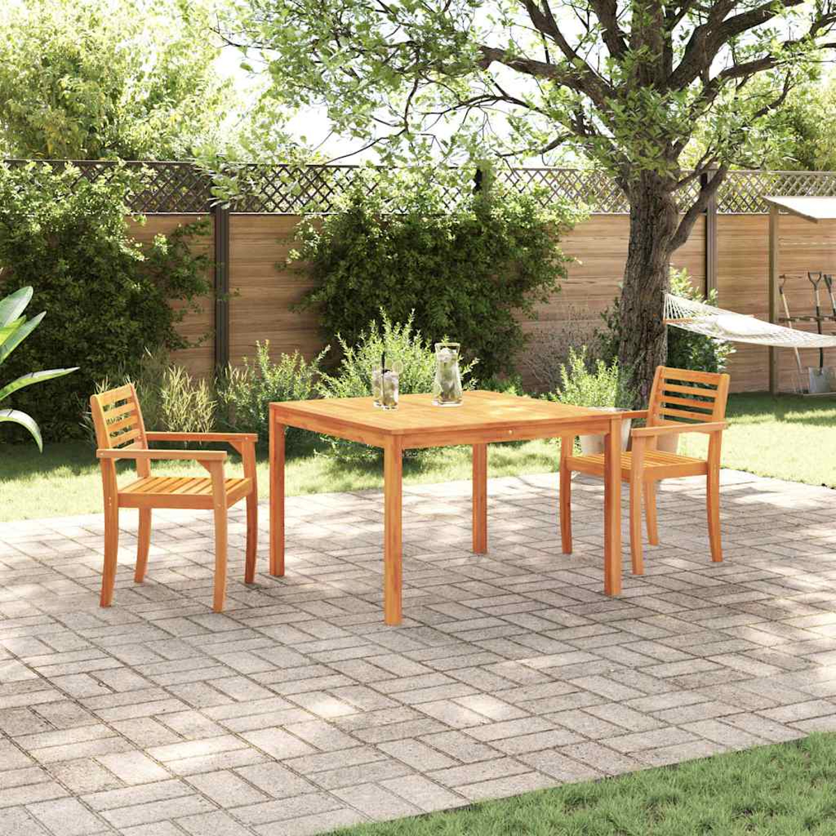 Mesa de madera con dos sillas en un jardín. Fondo con árboles, valla y hamaca. Dos vasos sobre la mesa.
