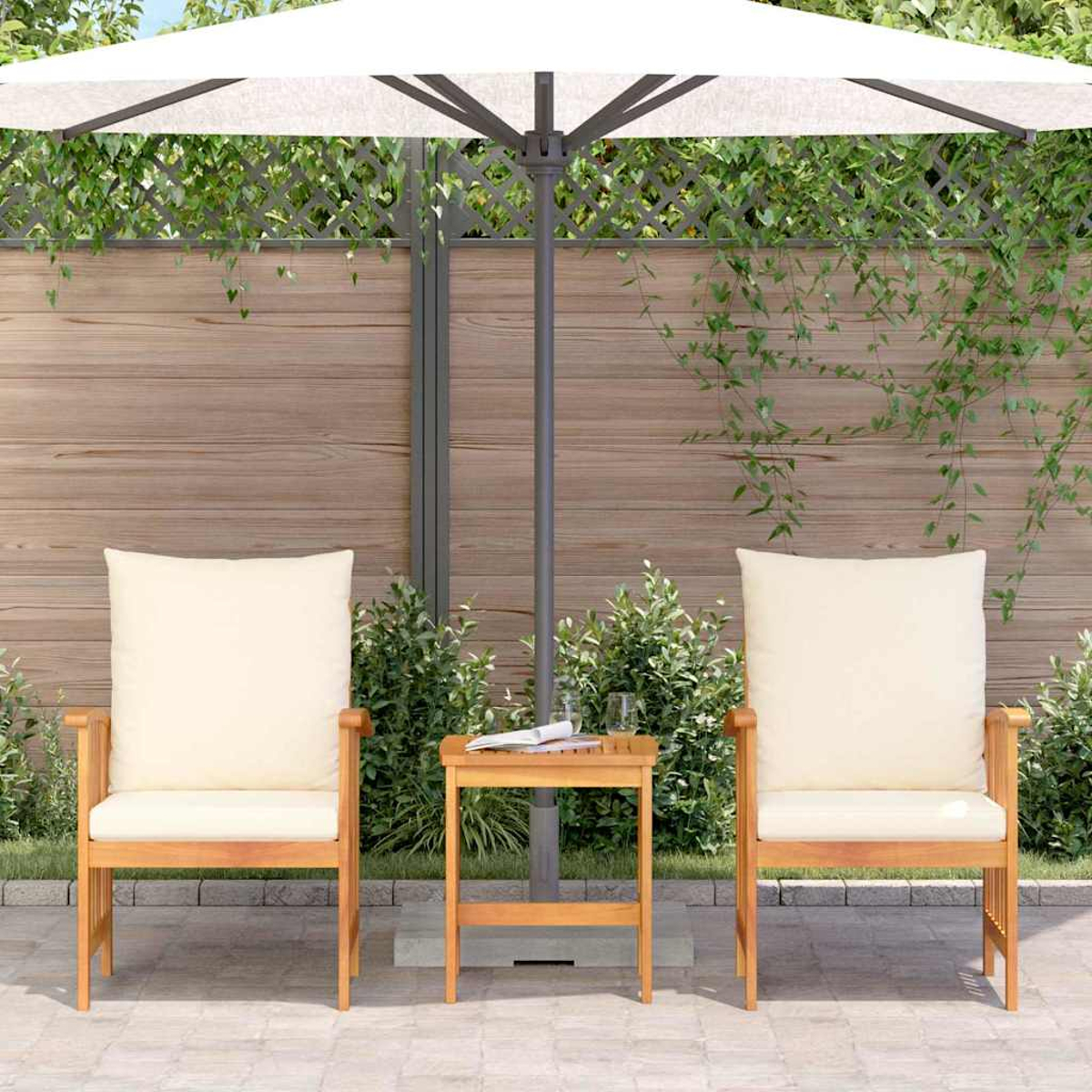 Dos sillas de madera con cojines y una mesa pequeña bajo una sombrilla blanca. El fondo es una valla de madera con plantas verdes.