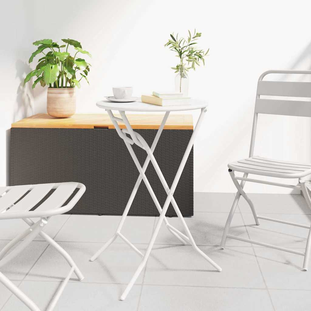 Weißer Tisch und Stühle auf einer Terrasse. Auf dem Tisch stehen eine Topfpflanze und eine Vase mit Blättern, im Hintergrund eine Truhe.